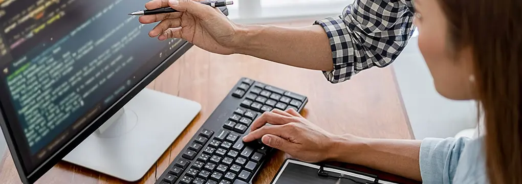 Hand pointing at code on a computer monitor while someone works on a keyboard.