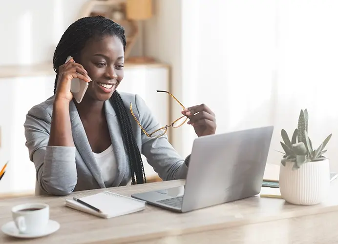 Woman holding eye glasses talking on phone in front of laptop.